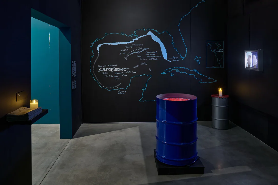 Gallery installation with a large blue crude-oil drum and video projection
